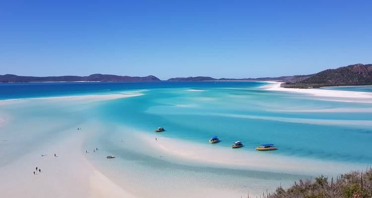 Aerial view of a stunning beach with clear turquoise waters and small boats, surrounded by lush hills.