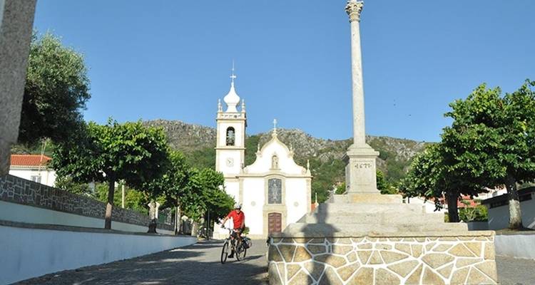 Een fietser pauzeert met zijn fiets voor een witte kerk en stenen kruis op een zonnig dorpsplein in Portugal.