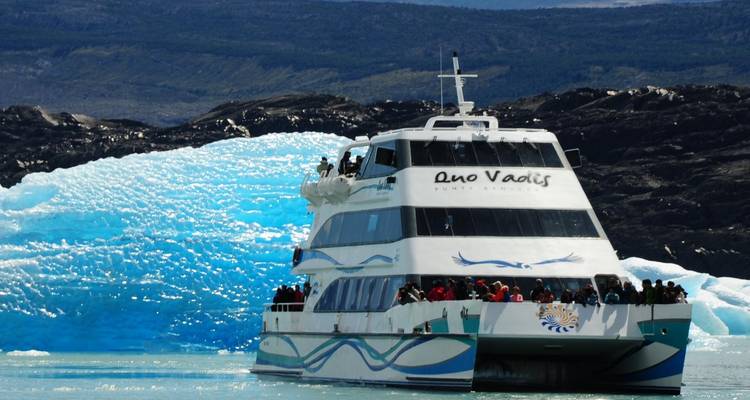 A boat full of people traveling near a large blue iceberg.