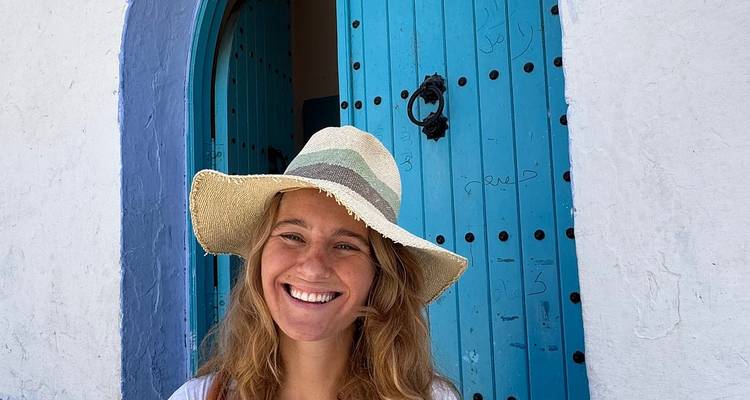 Une jeune femme coiffée d'un chapeau de paille sourit devant une porte voûtée d'un bleu éclatant à Chefchaouen.