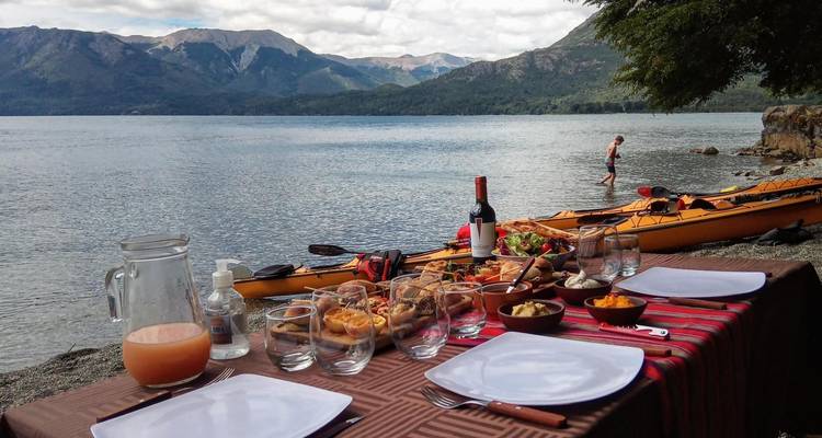 Configuración de comedor junto al lago con kayaks en el fondo, con vista a las montañas y el lago.