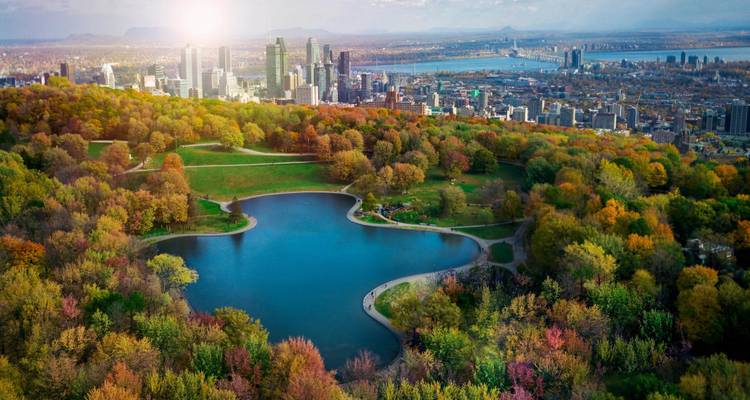 Vue aérienne d'un lac en forme de cœur dans un parc urbain forestier aux couleurs d'automne éclatantes, regardant vers la skyline moderne du centre-ville d'une grande ville bordée par un large fleuve sous un soleil brillant.