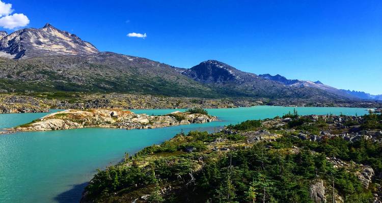Leuchtend türkisfarbener See, umgeben von felsigen Bergen und immergrünen Bäumen unter einem klaren blauen Himmel.