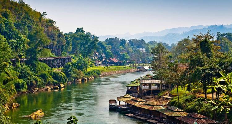 River with traditional wooden houses in a lush landscape in Thailand.