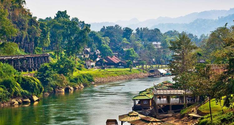 Des méandres de rivière verdoyante passent devant des pavillons en bois et le pont historique du chemin de fer Thaï-Birmanie dans les collines vertes.
