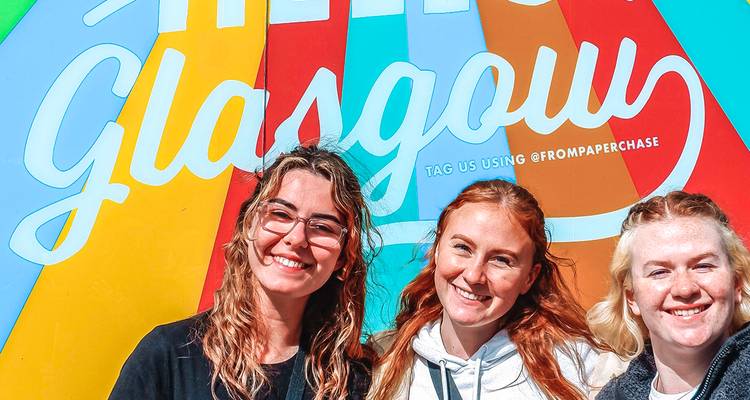 Tres mujeres jóvenes sonríen frente a un colorido mural callejero 'Hello Glasgow' bajo la brillante luz del día