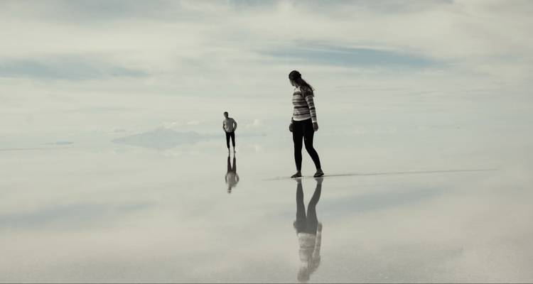 Deux voyageurs marchent sur un désert de sel aux surfaces miroir, leurs reflets flottant dans le ciel brumeux.