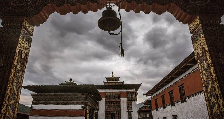 Cour de temple bhoutanais encadrée par une arche ornementée et une cloche suspendue sous des nuages gris dramatiques.