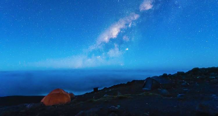 Cielo nocturno lleno de estrellas con la Vía Láctea arqueándose sobre las tiendas de campaña en un campamento de montaña.