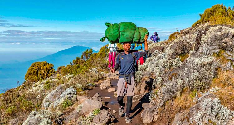 Porter equilibra una gran bolsa verde sobre su cabeza mientras camina por un sendero alpino.