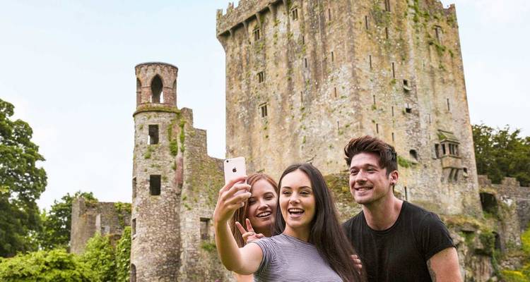 Tres amigos se toman una selfie frente a las rugosas paredes de piedra del Castillo de Blarney cubiertas de hiedra.