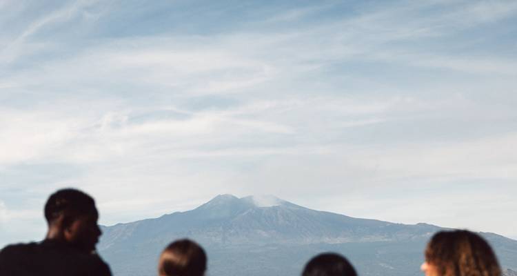 Visitantes en silueta admiran el humeante Monte Etna bajo un cielo azul despejado.