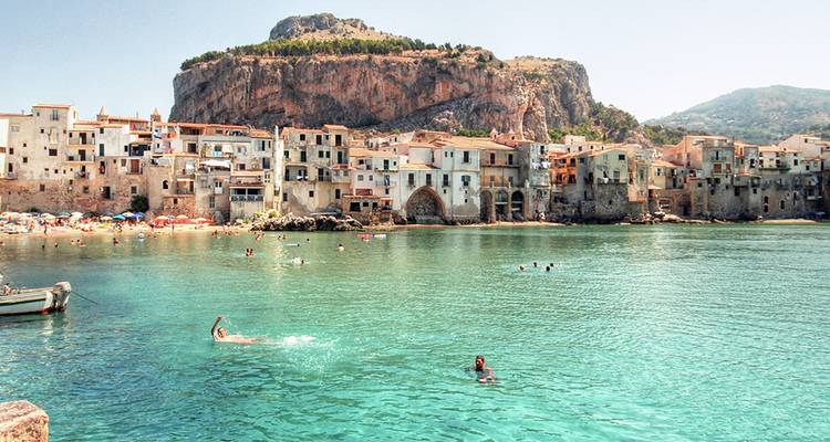 Village côtier historique adossé à une falaise escarpée, eau turquoise au premier plan avec plusieurs nageurs profitant de la baie.