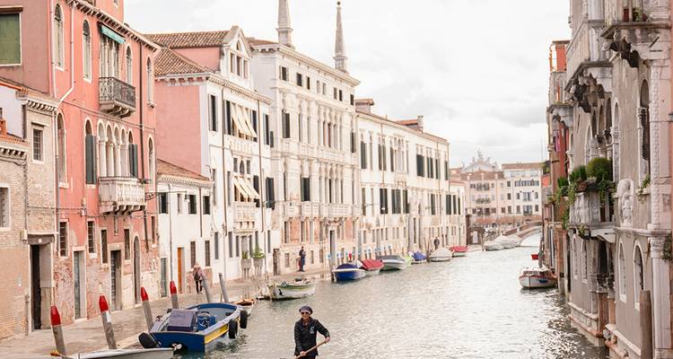 Un gondolero rema por un tranquilo canal veneciano bordeado de palacios de colores pastel y botes amarrados en un día soleado
