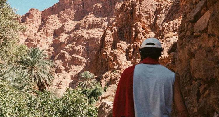 Vista trasera de un senderista con gorra y bufanda roja mirando hacia arriba en dirección a los imponentes acantilados rojos y las palmas de oasis en la Garganta de Todra.