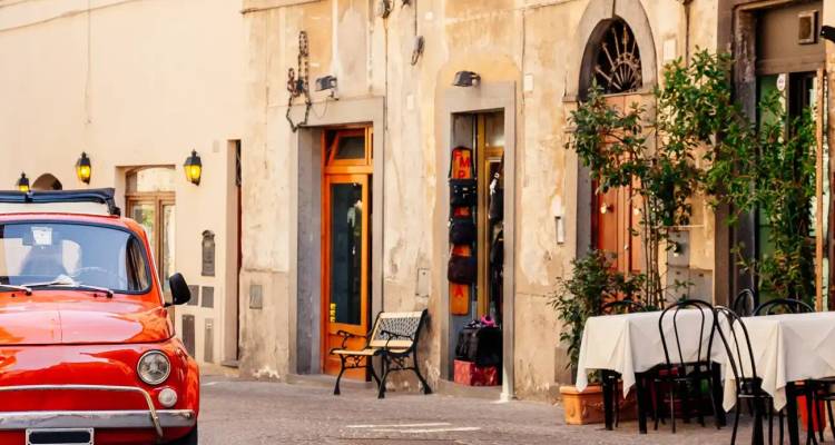 A classic red Fiat and café table decorate a charming narrow street lined with aged stone buildings.