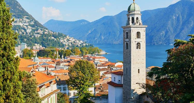 A scenic view of Lugano with a lake, bell tower, and surrounding hills.