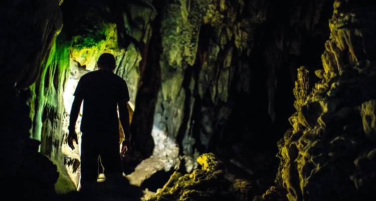 Un aventurier en silhouette explore une grotte de calcaire sombre illuminée par une lumière naturelle verte.