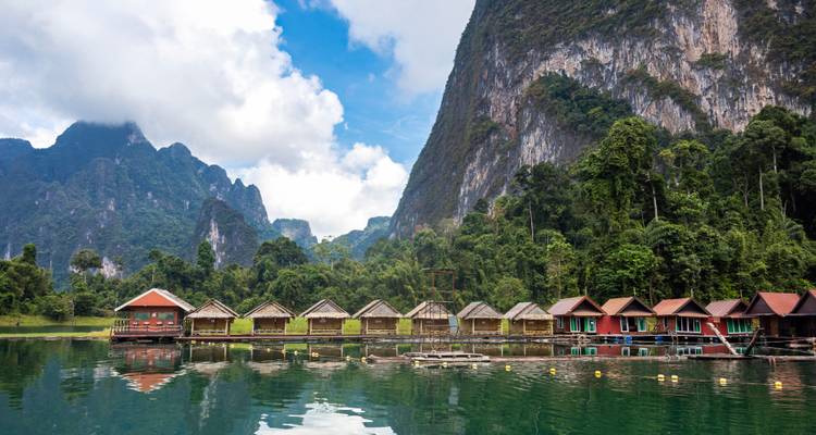Rangée de bungalows flottants sur un lac aux eaux cristallines entouré de falaises calcaires imposantes recouvertes de jungle.