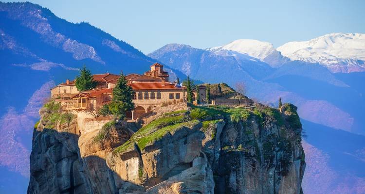 A monastery balances dramatically on a towering rock pillar with snow-capped mountains in the distance.