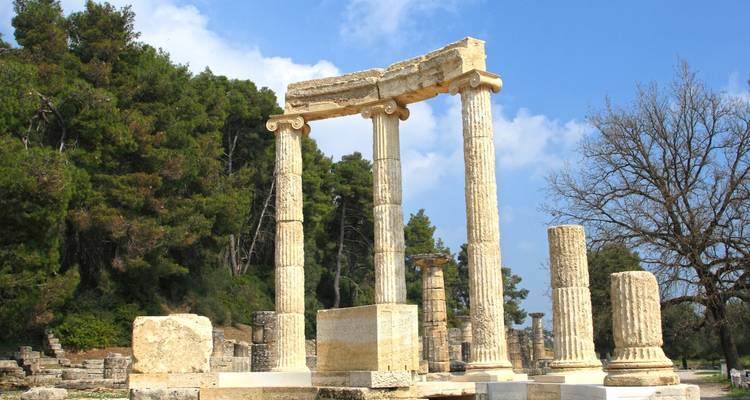 Ancient stone columns and ruins stand against a backdrop of pines and blue sky.