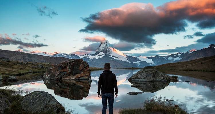 Homme debout au bord d'un lac avec vue sur une montagne enneigée.