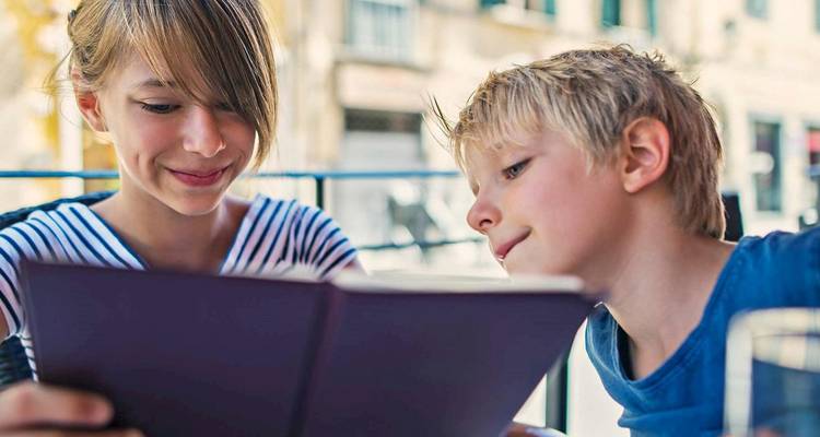 Dos niños se sientan en una mesa de café al aire libre estudiando un menú bajo una suave luz del día.