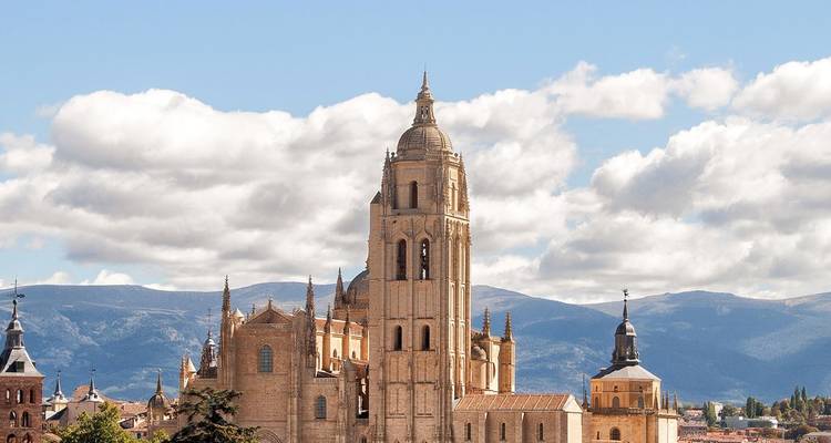 Majestuosa Catedral de Segovia elevándose sobre tejados de tejas con montañas de fondo y nubes esponjosas.