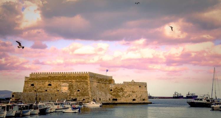 Forteresse historique avec un ciel rose et des bateaux au premier plan.