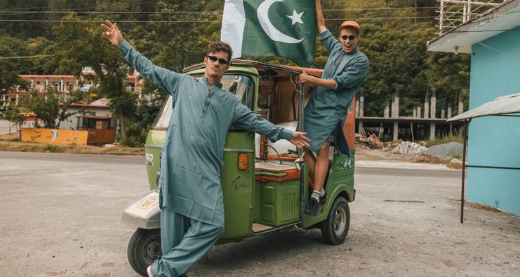 Twee reizigers in traditionele shalwar kameez poseren energiek met een groene tuk-tuk en een grote Pakistaanse vlag in een bergachtige straatse omgeving.