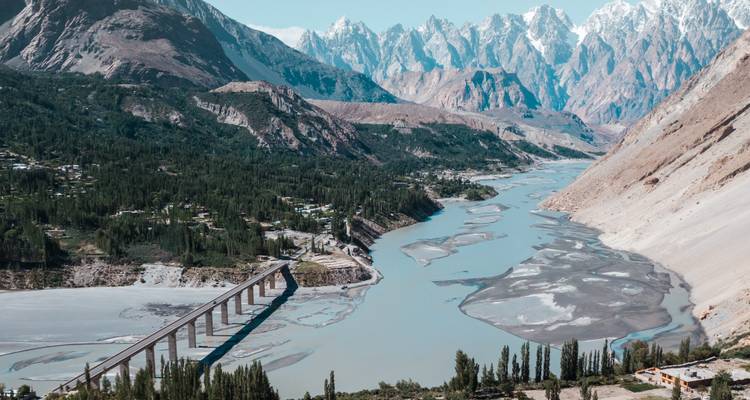 Breed panoramisch uitzicht op een hangbrug die een gevlochten turquoise rivier oversteekt onder het dramatische Karakoram-gebergte.