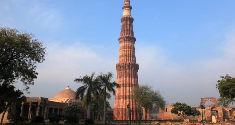 De Qutub Minar omringd door bomen en ruïnes in een parkachtige omgeving.