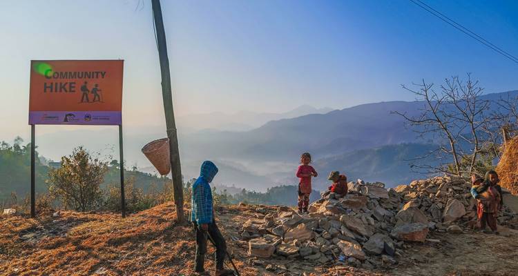 Children standing near a ‘Community Hike’ sign on a hillside trail with hazy mountain layers at dawn