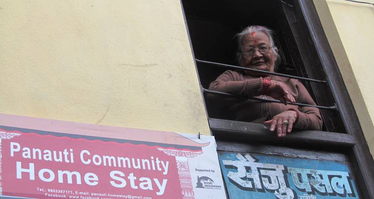 Elderly woman smiles from a window above a Panauti Community Home Stay signboard