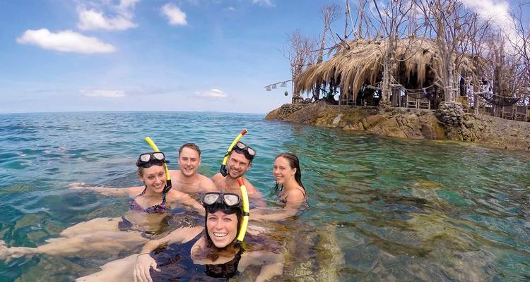Grupo de personas haciendo snorkel cerca de una playa tropical con una cabaña de techo de paja en el fondo.