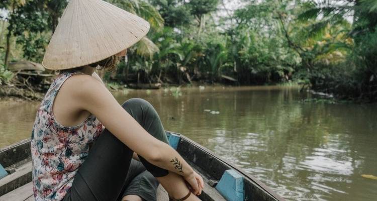 Una joven mujer que usa un sombrero cónico se sienta en la proa de un bote de madera mientras se mueve a lo largo de un río angosto rodeado de selva.
