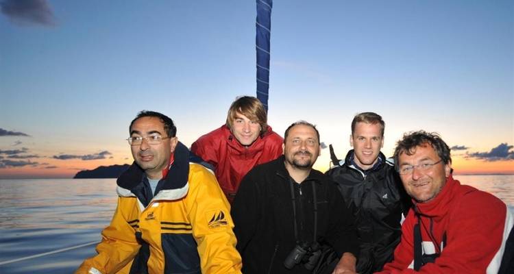 Cinco personas vestidas con chaquetas sonriendo a bordo de un barco al atardecer.