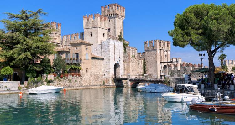 Medieval lakeside castle with crenellated towers reflected in calm turquoise water alongside moored boats.