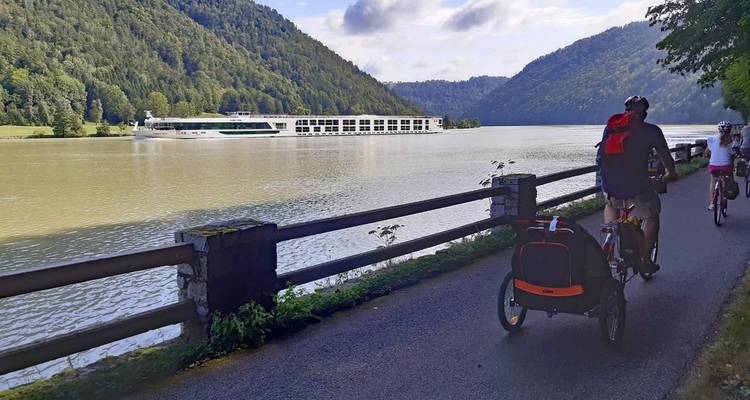 Radfahrer auf einem Weg an einem Fluss mit einem Kreuzfahrtschiff im Hintergrund.