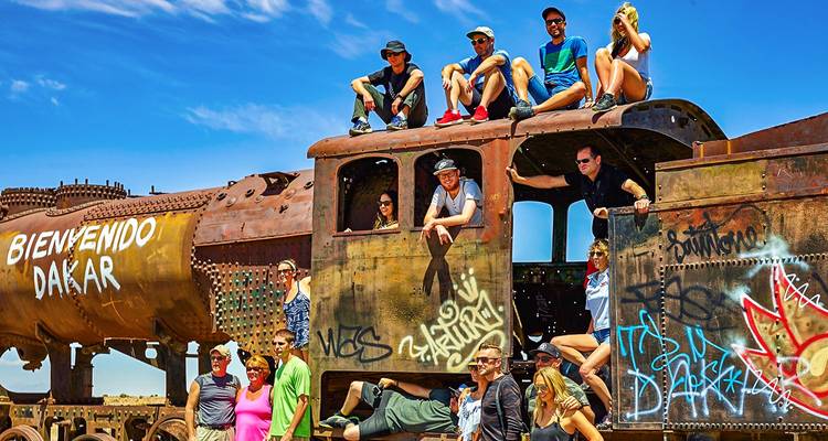 Eine lebhafte Gruppe posiert auf einer verrosteten Dampflokomotive, die mit Graffiti besprüht ist, auf dem Zugfriedhof von Uyuni unter einem leuchtend blauen Himmel.