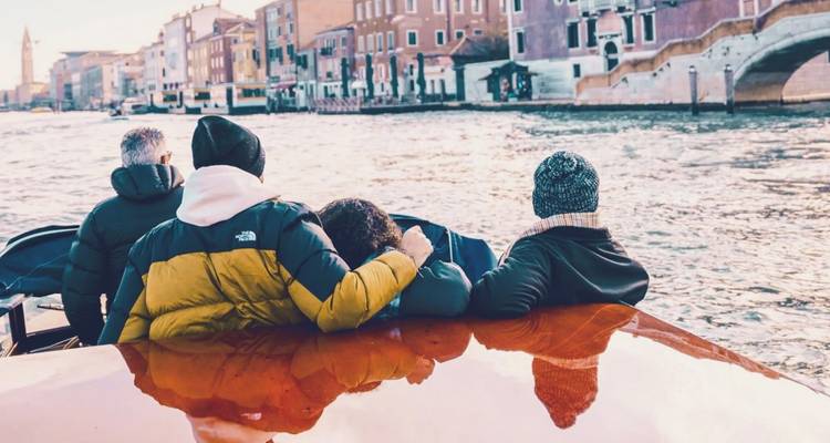 Quatre personnes sont assises dans un taxi nautique en bois qui navigue dans un canal vénitien bordé de bâtiments historiques aux couleurs pastel à la lumière du coucher de soleil.