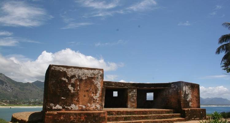 Oud stenen fort met uitzicht op een strand met verre bergen en blauwe lucht.