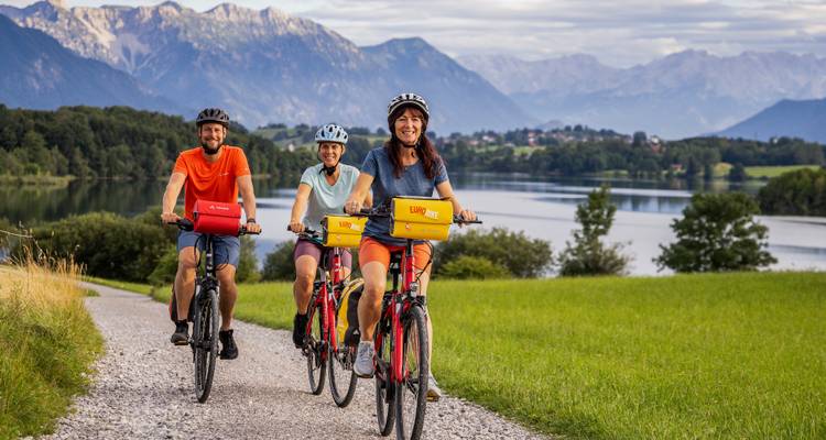 Trois cyclistes souriants font du vélo électrique sur un sentier en gravier au bord d'un lac avec des paysages alpins et des montagnes en couches à l'arrière-plan.