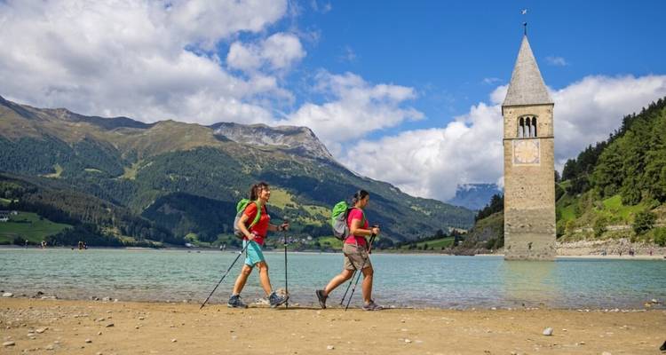 Wanderer, die am Seeufer entlanggehen, mit einem Glockenturm, der aus dem Wasser ragt.