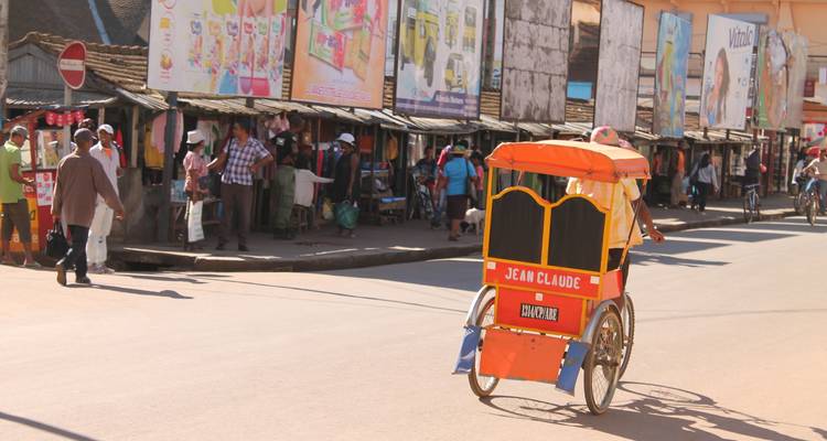 Un homme conduisant un véhicule coloré à trois roues dans une rue avec des étals de marché et des panneaux publicitaires.