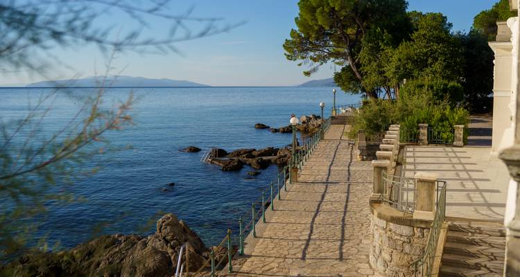 Promenade côtière en pierre avec rambardes et arbres surplombant les eaux calmes et bleues de la mer Adriatique ; une personne seule contemple les îles lointaines dans une lumière chaleureuse.