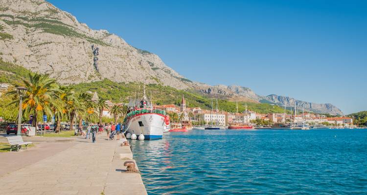 Promenade portuaire vibrante avec palmiers, un bateau d'excursion orné et une mer turquoise éclatante se dressant contre d'imposantes montagnes rocheuses et une ville côtière.