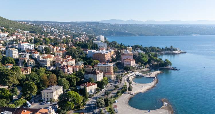 Vue aérienne d'une ville adriatique en montagne avec des villas historiques et des hôtels longeant la côte, une promenade sinueuse et des plateformes de baignade à côté d'une eau bleu profond.