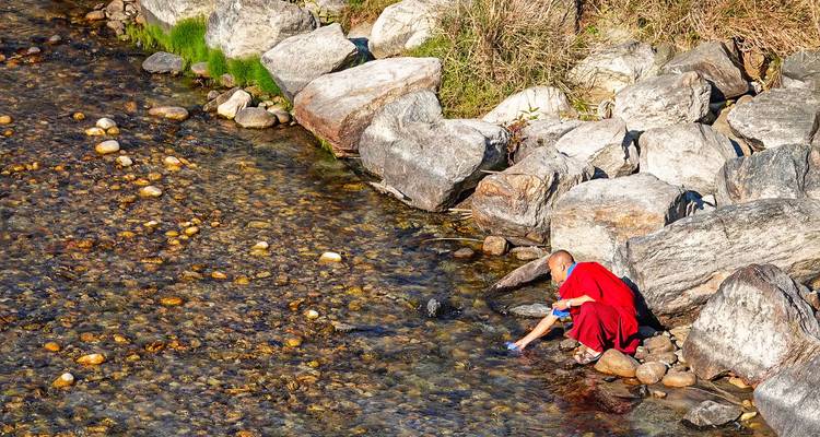 Monnik in rode gewaden gehurkt naast een rivier met rotsen.
