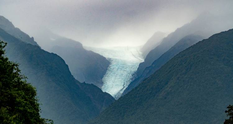 Lengua glacial brumosa extendiéndose entre montañas verdes escarpadas bajo nubes bajas.